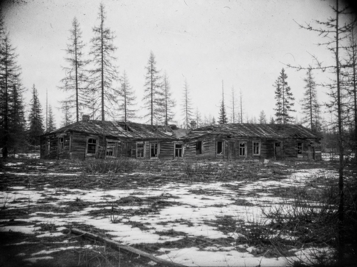 A grainy black-and-white photograph of a long collapsed wooden barracks among larch trees, patches of snow on the ground.