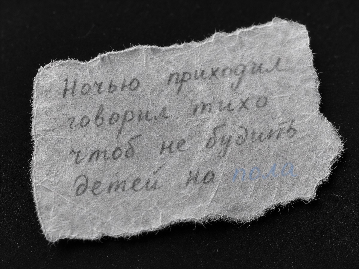 A macro photograph of a torn scrap of cigarette paper on a black surface, four lines of faint pencil handwriting in Russian; the final word carries a faint blue tint in an otherwise black-and-white photograph.